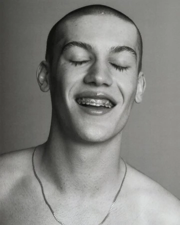 Monochrome portrait of a young adult male with closed eyes, smiling and wearing braces and a chain necklace against a neutral background.