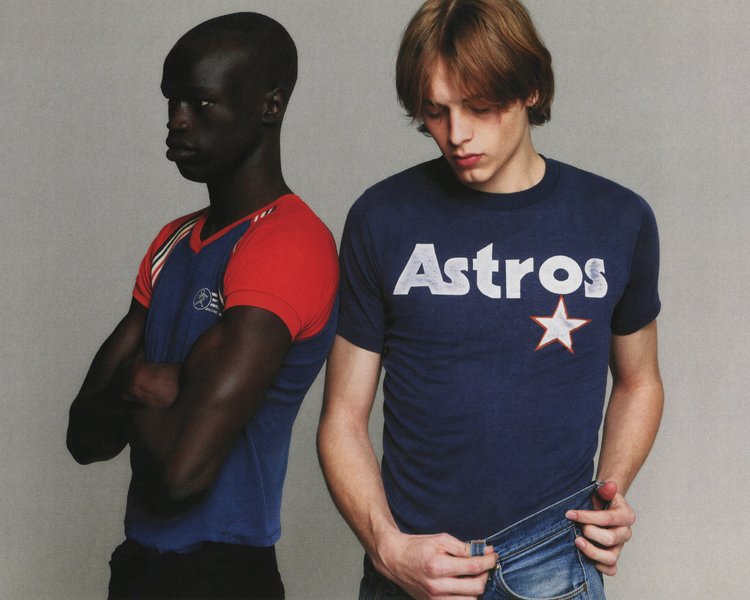 Two adult men pose in t-shirts and jeans against a neutral background, showcasing casual fashion with an editorial style.
