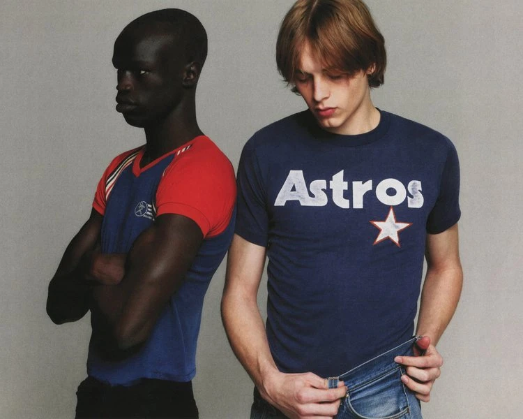 Two adult men pose in t-shirts and jeans against a neutral background, showcasing casual fashion with an editorial style.