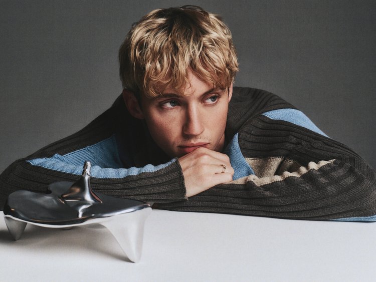 Portrait of a blond adult with a contemplative expression, resting head on a metallic object and wearing a brown and blue sweater.