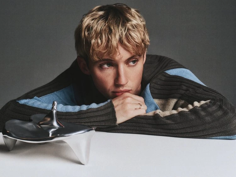 Portrait of a blond adult with a contemplative expression, resting head on a metallic object and wearing a brown and blue sweater.