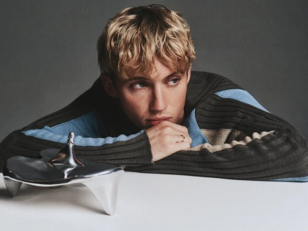 Portrait of a blond adult with a contemplative expression, resting head on a metallic object and wearing a brown and blue sweater.