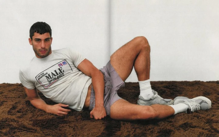 A man wearing a printed t-shirt and patterned shorts lies on a dark brown shaggy carpet against a white wall.