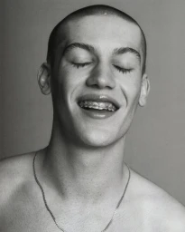 Monochrome portrait of a young man with a shaved head and dental braces smiling with eyes closed, wearing a thin chain necklace.
