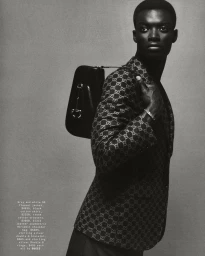Black man in a patterned monogram blazer carrying a black leather shoulder bag against a grey backdrop, photographed by Joe Brennan.