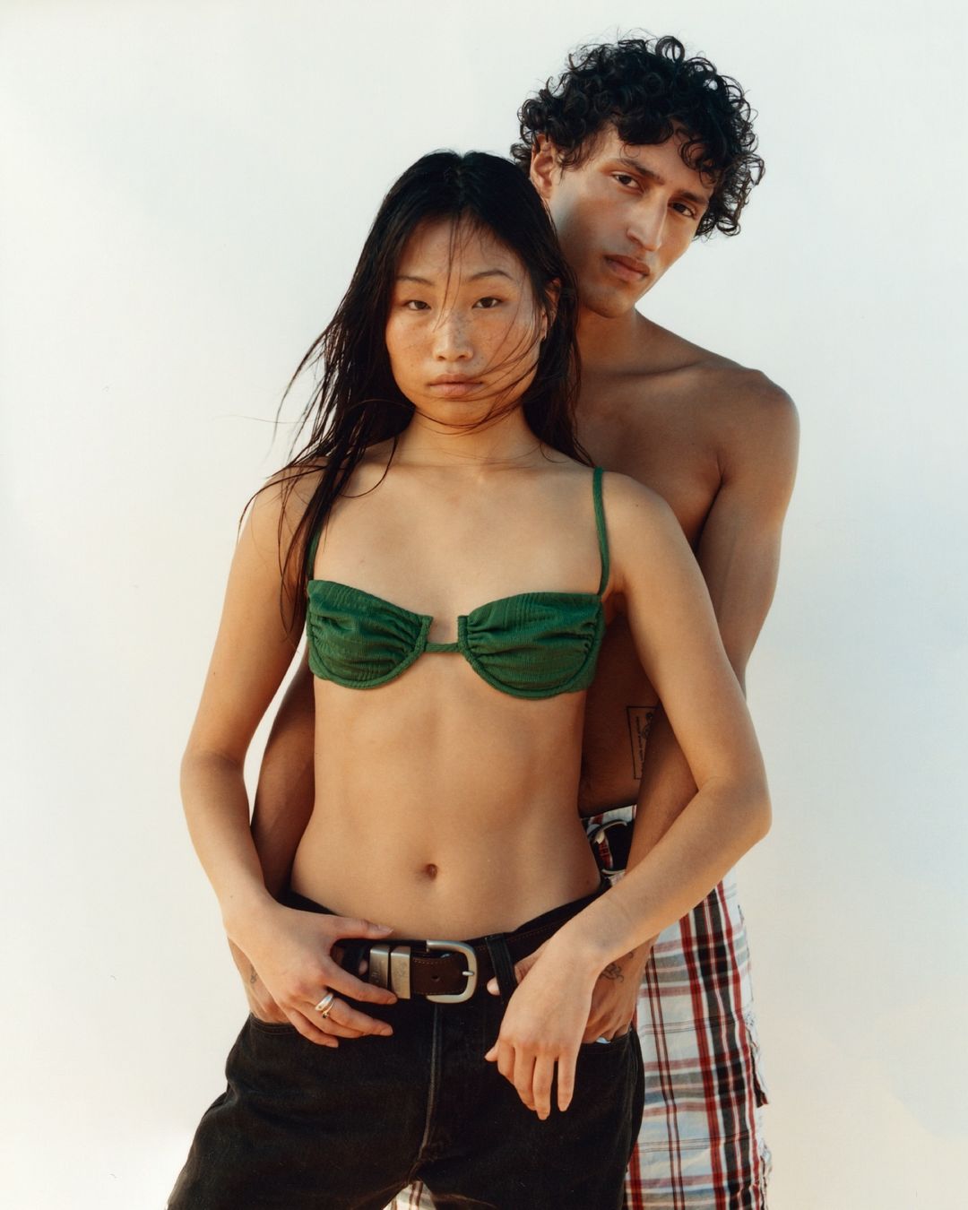 An Asian woman in a green bikini top poses with a curly-haired man against a white backdrop in a fashion editorial shot.