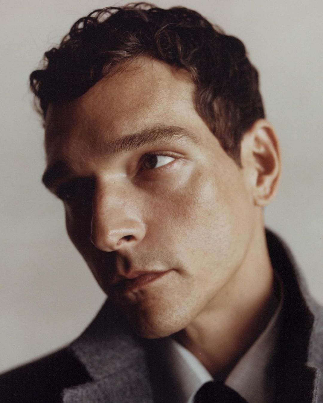Close-up portrait of an adult male with dark curly hair wearing a grey suit and tie, against a light grey backdrop in a photo studio.