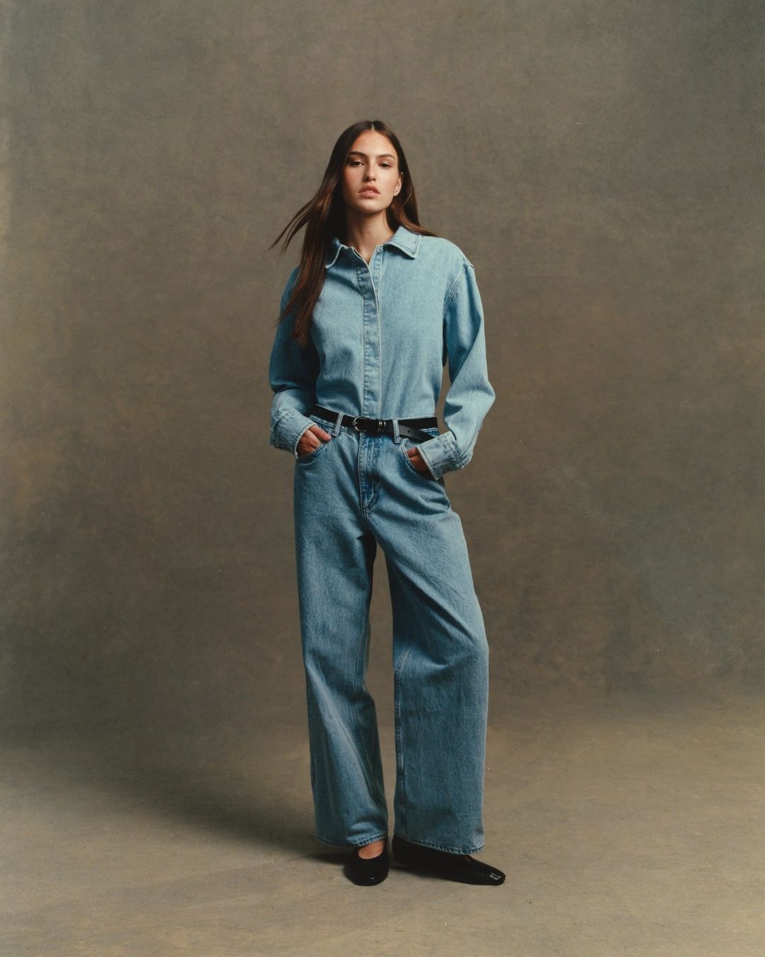 Fashion shot of a woman in denim shirt and flared jeans, hands in pockets, black belt and flats, studio background, natural light.