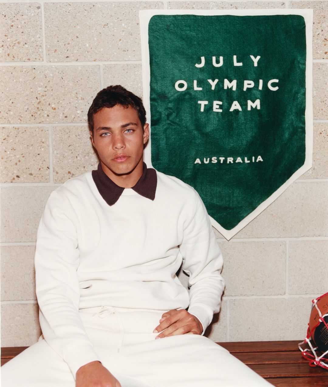 A young, light-skinned male wearing a vintage white pullover with a brown collar poses indoors with an Olympic banner in the background.