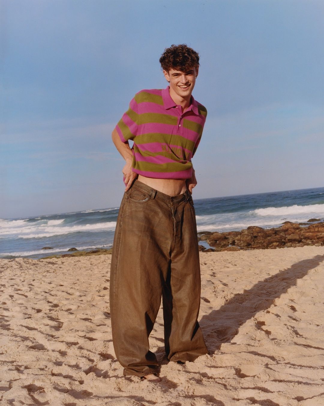 Young adult male poses on a beach with olive and pink striped polo shirt and very large, baggy brown pants. Ocean and sky background.