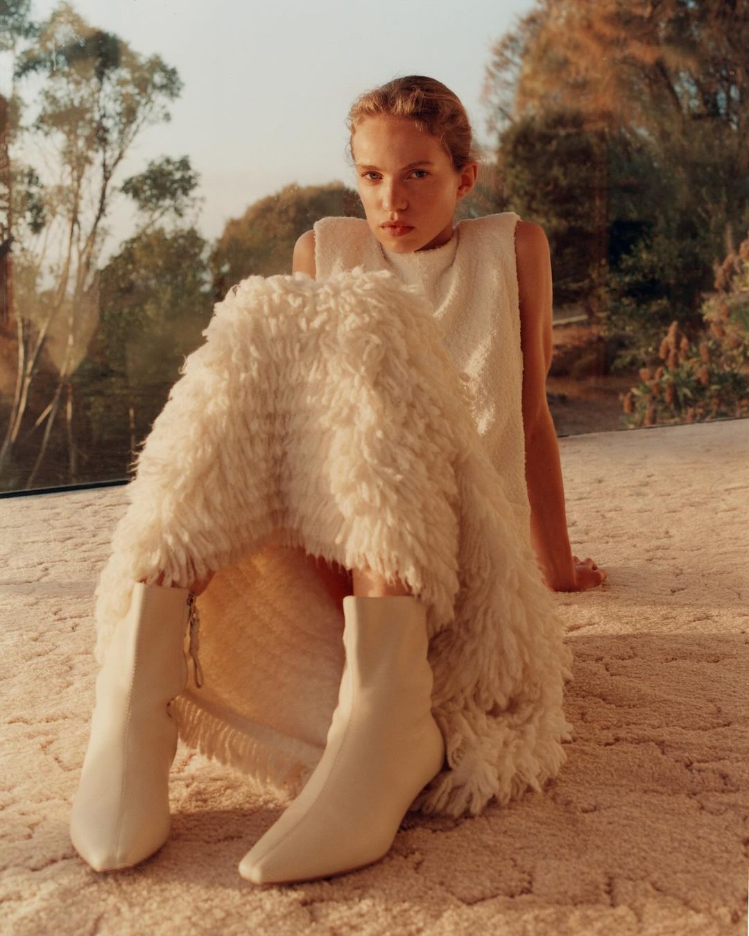 Fashion shot of a woman in a white dress and boots sitting on a textured floor, with natural light and trees visible in the background.