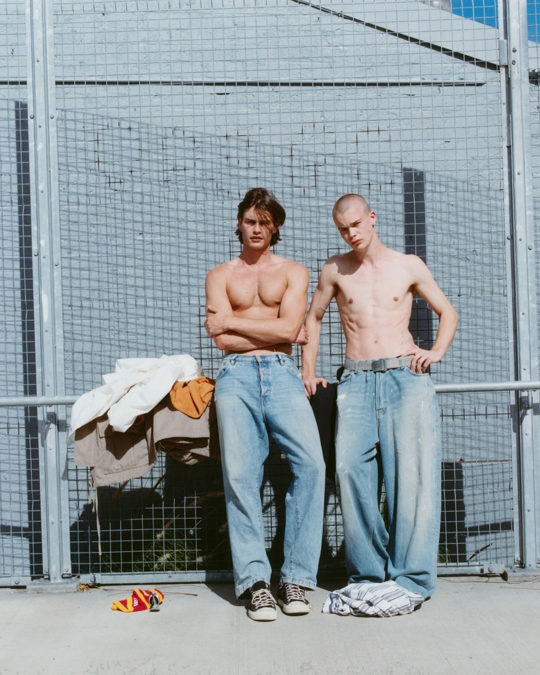 Two shirtless young adult men pose in baggy jeans outdoors in front of a wire fence on a sunny day. Fashion and style.