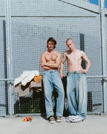 Two shirtless young adult men pose in baggy jeans outdoors in front of a wire fence on a sunny day. Fashion and style.
