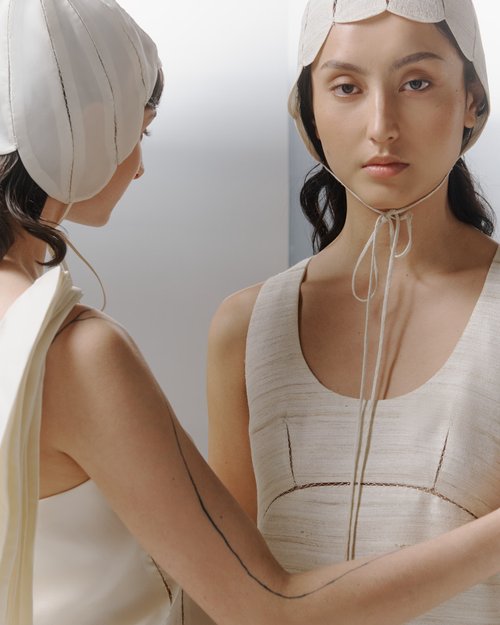 Two women pose in matching cream-colored linen dresses and head coverings in a minimalist studio setting with soft, neutral lighting.