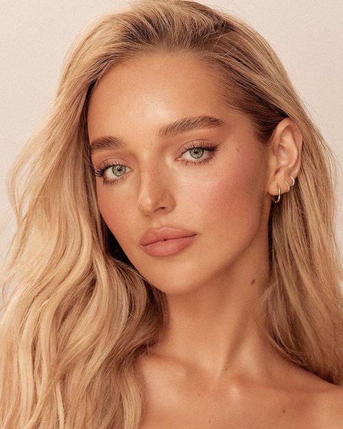 Close-up portrait of a blonde woman with fair skin, light eyes, and subtle makeup against a light background. She wears hoop earrings.
