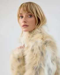 Close-up portrait of a blonde woman wearing a fluffy cream faux fur coat against a solid white background.