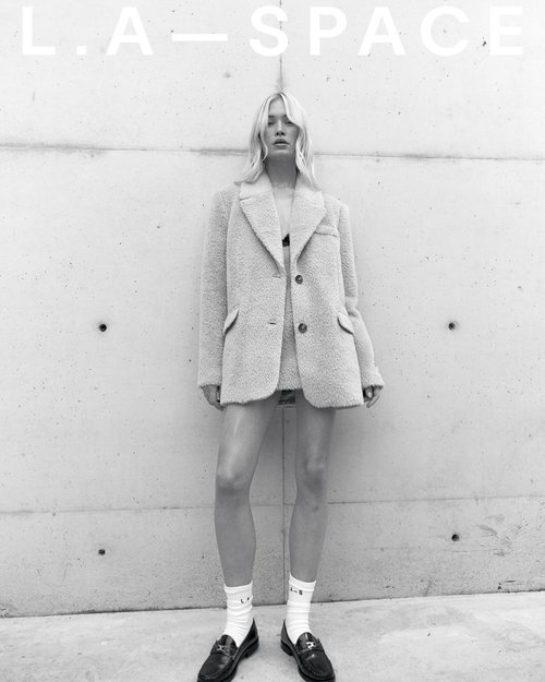 A slender fashion model in a wool-like blazer and skirt stands in front of a concrete wall wearing socks and loafers in a studio shot.