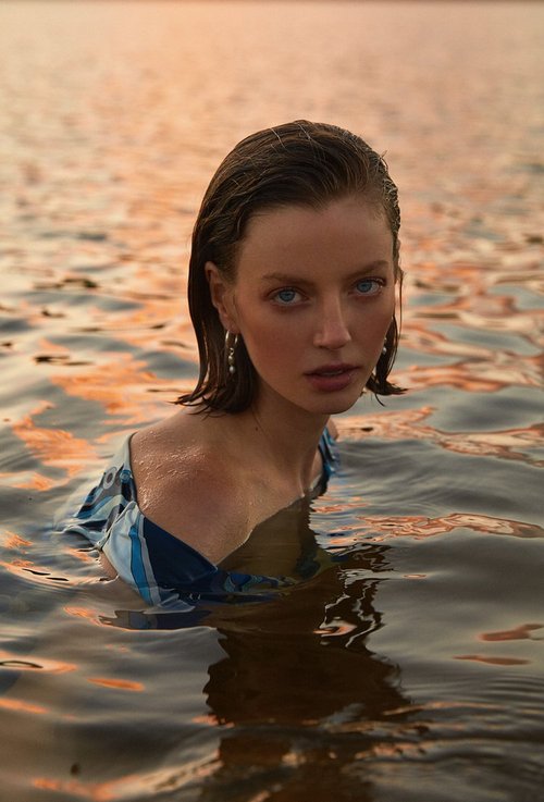 Close-up portrait of a young woman waist-deep in water with a blue and white patterned top, wet hair, and pearl earrings at sunset.