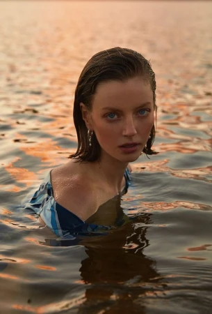 Close-up portrait of a young woman waist-deep in water with a blue and white patterned top, wet hair, and pearl earrings at sunset.