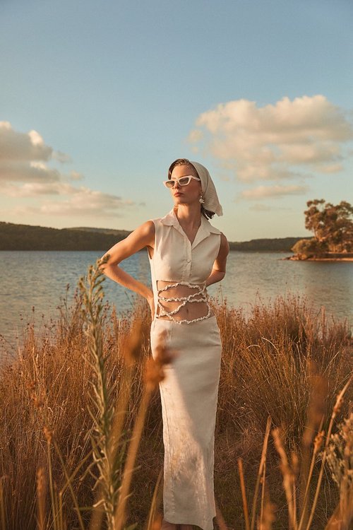Woman in cream dress, scarf, and sunglasses poses outdoors against a lakeside backdrop at golden hour, showcasing fashion and style.