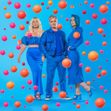 Three people (one man, two women) in blue outfits posing in a studio with pink and orange floating spheres, creating a surreal and colorful composition.