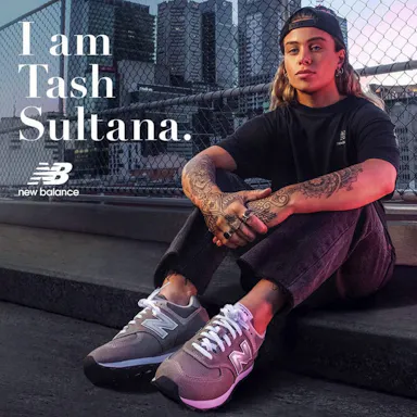 Tash Sultana wearing New Balance sneakers, sitting on concrete stairs in a city environment with urban buildings in the background.