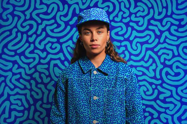 Portrait of a young adult woman wearing a matching blue patterned cap and jacket against a blue patterned backdrop, smiling and looking at the camera.