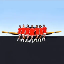 Seven men in matching orange t-shirts and white shorts posing in front of a yellow airplane against a blue sky