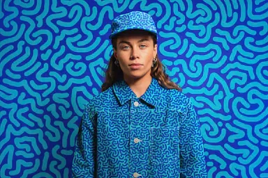Portrait of a person in a blue patterned jacket and matching cap against a vibrant blue squiggly patterned background.