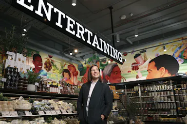Overhead shot of a woman standing in a grocery store beneath an 'Entertaining' sign surrounded by produce and shelved items.