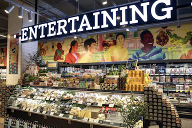 Overhead shot of a supermarket deli counter filled with food products under a large ‘Entertaining’ sign, with an illustration behind.
