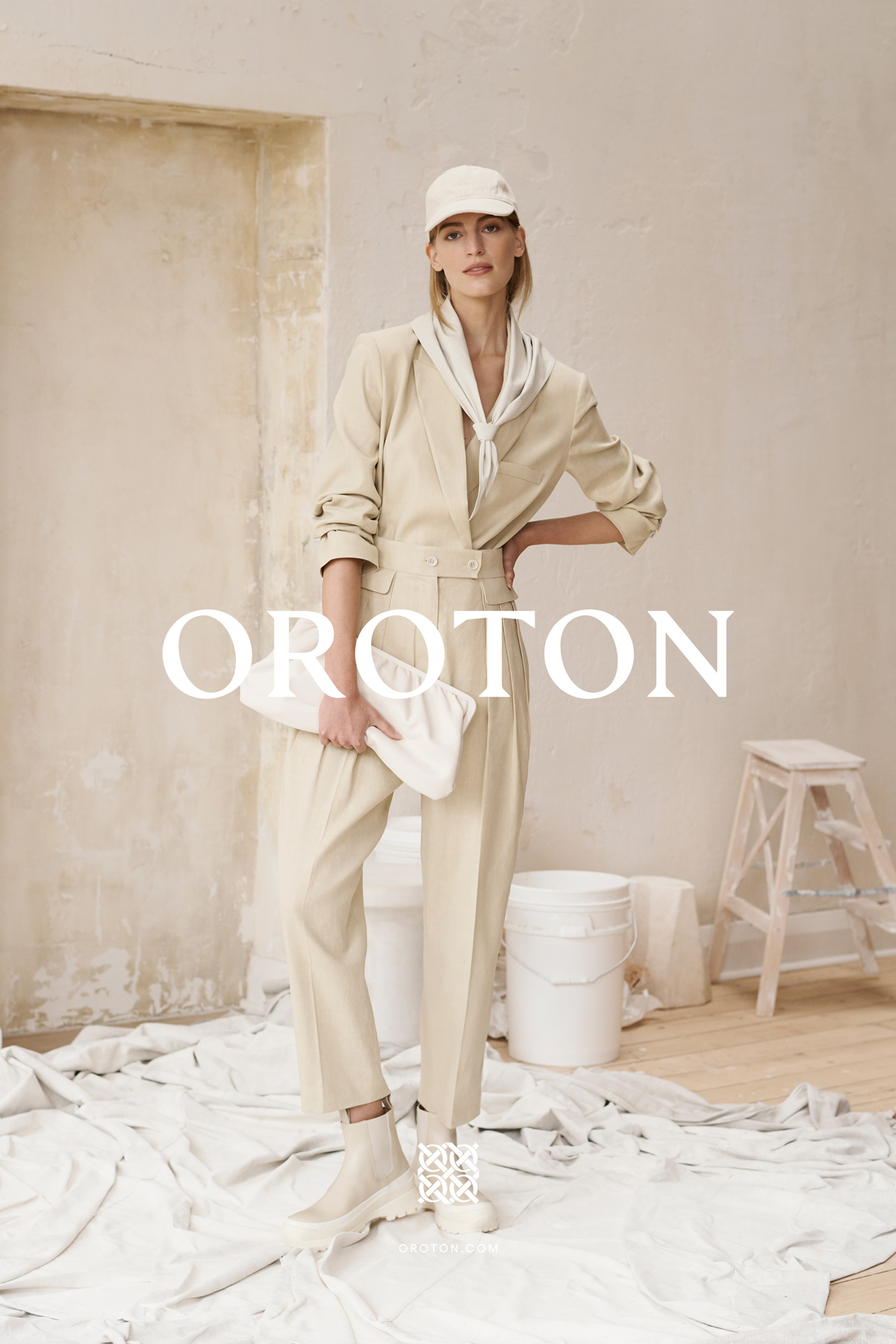 Fashion shot of an adult woman wearing a beige pantsuit and matching hat, holding a white clutch, in a studio with painting props.