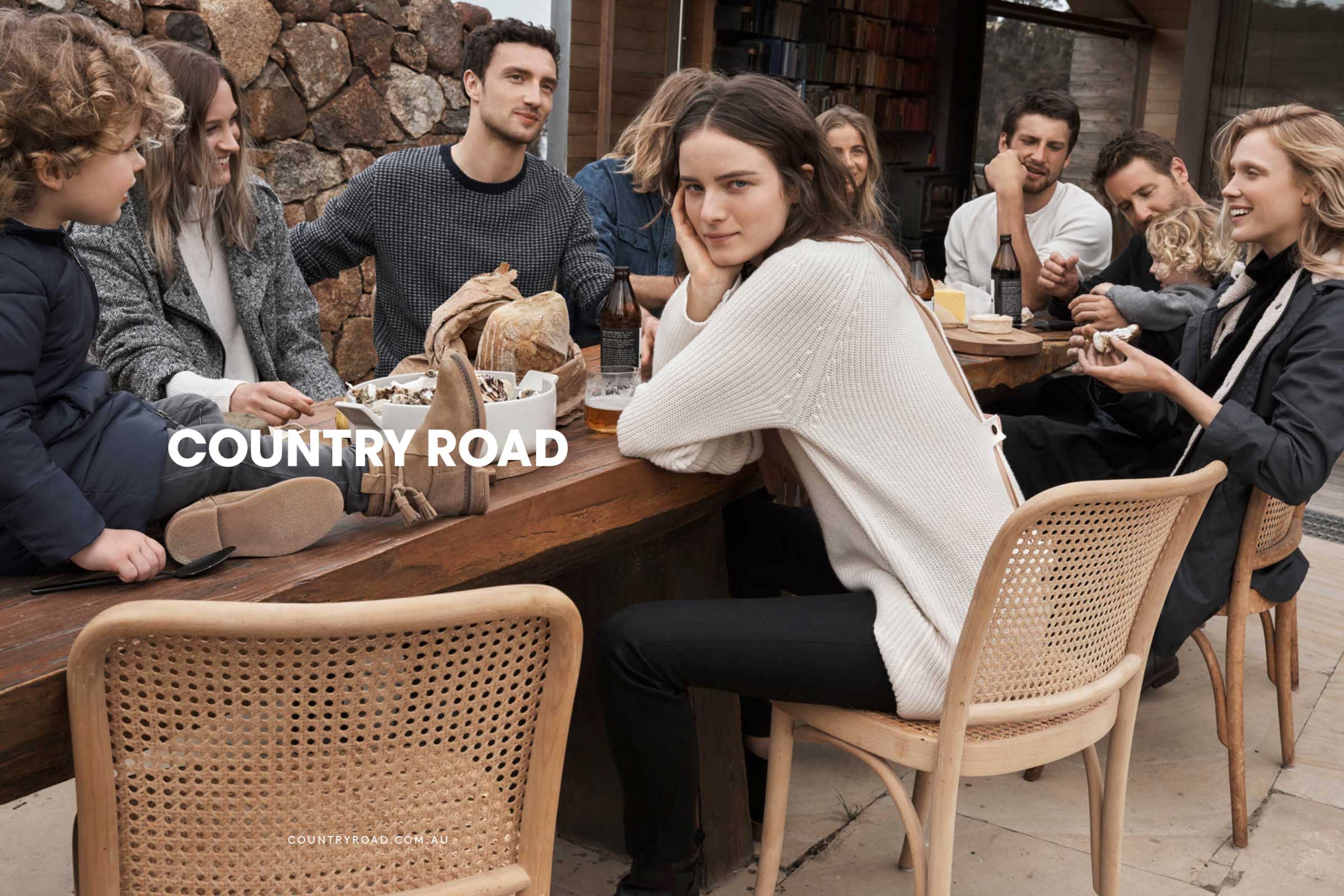 Group of people sitting at a wooden table outdoors, sharing food and drinks in natural daylight, stone wall and greenery background.