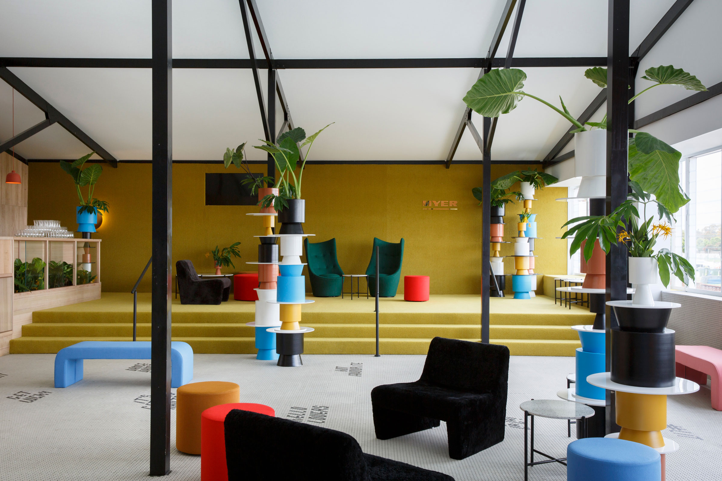 Eclectic modern interior design features green & black chairs, colorful stools, tiered pot plants, and black structural support columns.