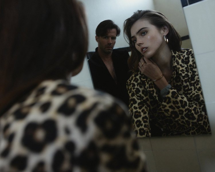 Woman in a leopard-print blazer looks into a mirror while a man stands behind her. Indoor fashion shoot with neutral colors.
