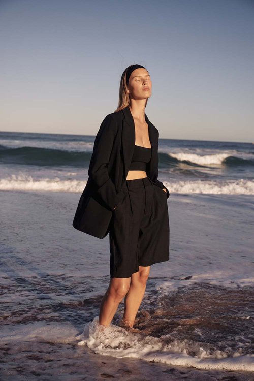 Woman in black blazer, top, and shorts stands in ocean surf on a sunny beach, eyes closed, looking up. Sky and waves background.