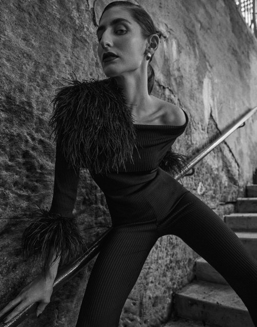 A striking black and white fashion shot of a woman posing in a feathered outfit on stairs, set against a textured stone wall backdrop.