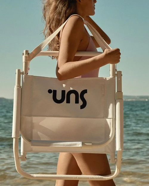 Woman carrying a folded white beach chair with black text against an ocean and sky background
