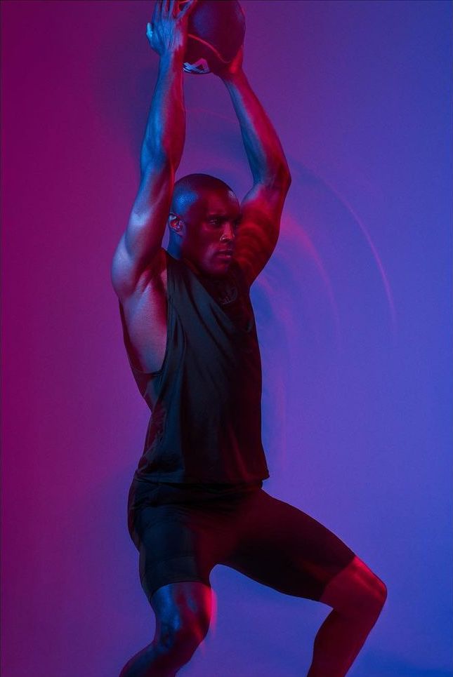 Athletic man in black activewear powerfully holds a medicine ball overhead with red and blue lighting and blurred motion in a studio setting.