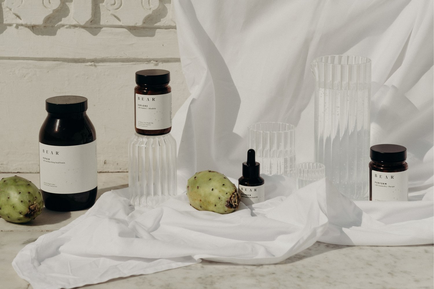Still life photo of BEAR brand supplement bottles and cacti fruits on a white draped fabric, set against a white wall.
