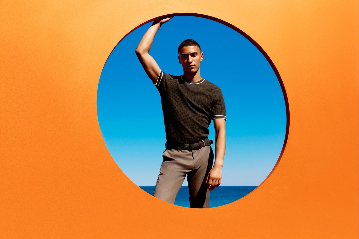 A young adult man poses in front of a circular orange frame, set against a bright blue sky and ocean backdrop on a sunny day.