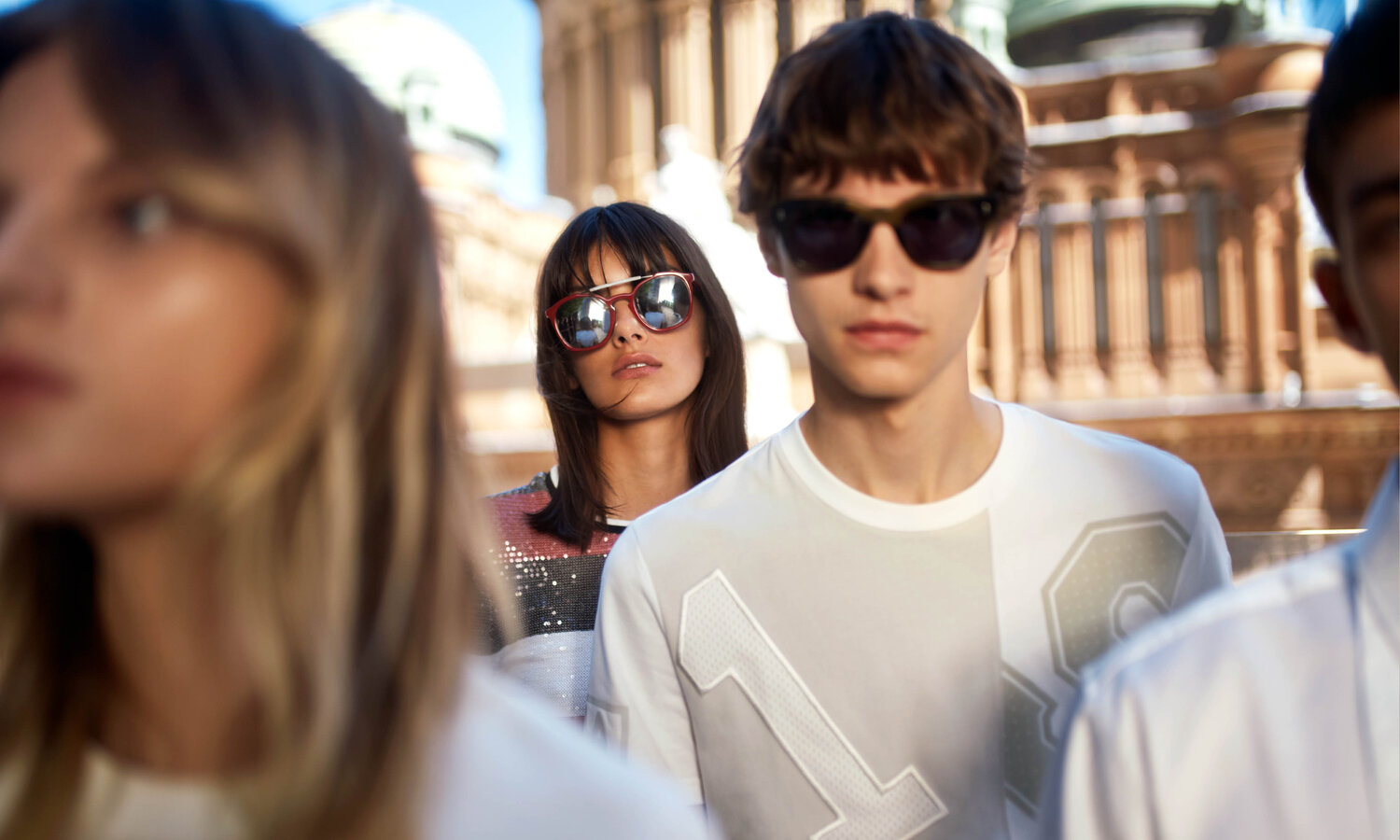 Group of young adults outdoors wearing sunglasses and casual clothing, with urban architecture visible in the background.