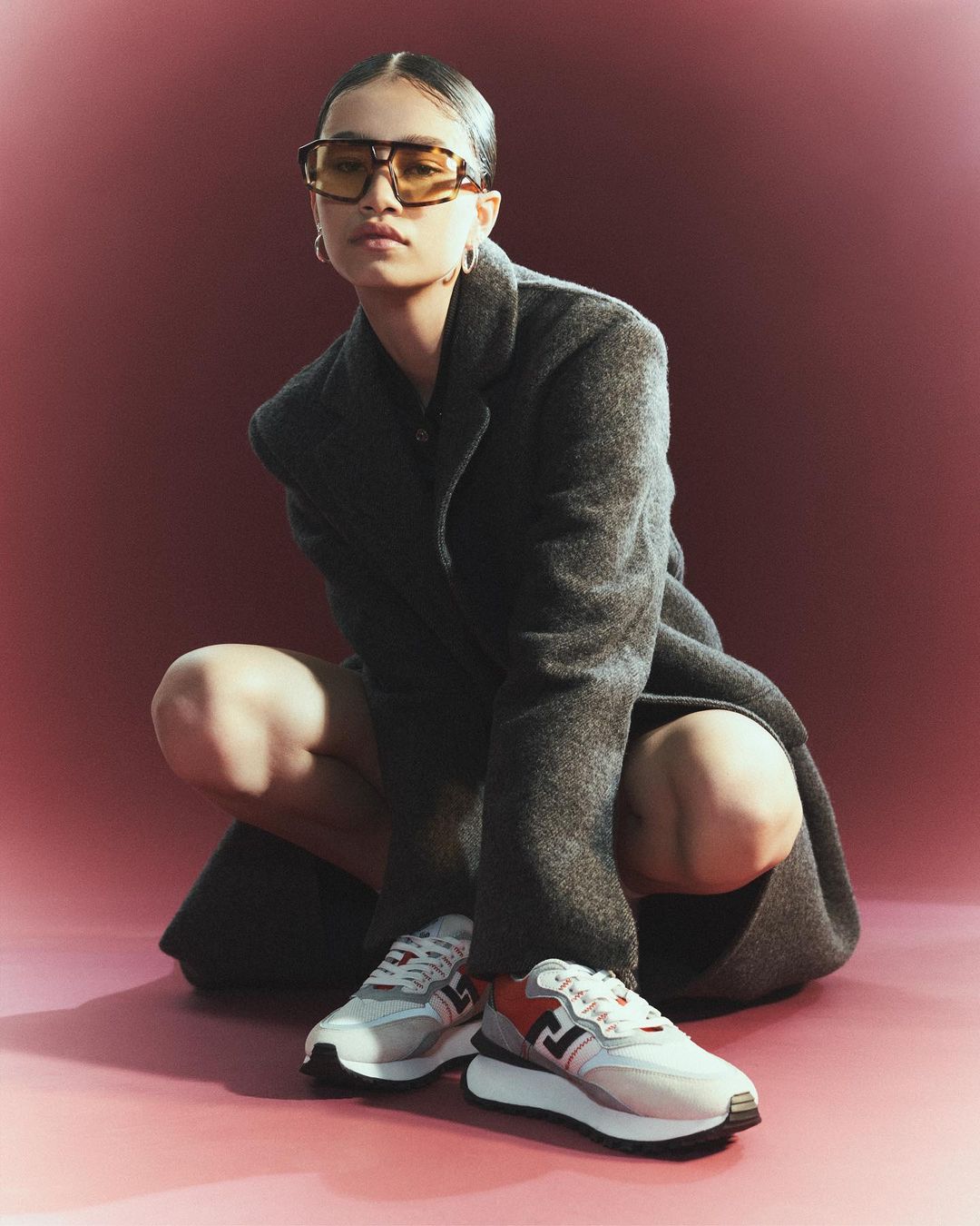 A person wearing a gray coat and yellow sunglasses is crouching and posing in front of a reddish background next to a pair of sneakers.