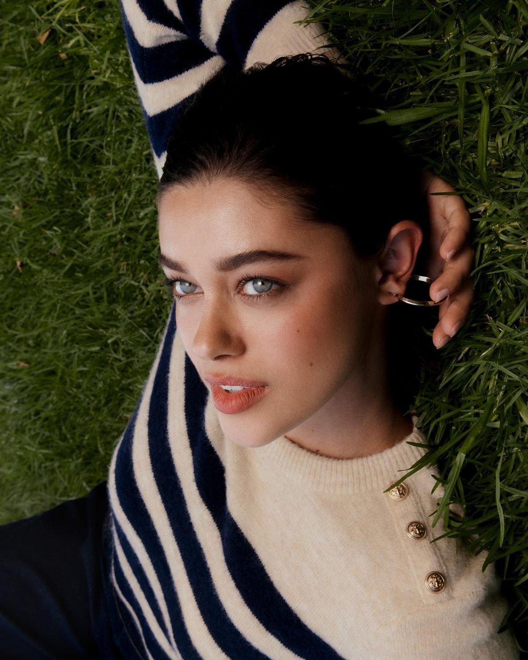 Portrait of a young woman lying on green grass, wearing a striped sweater, silver hoop earrings, and soft, natural makeup.