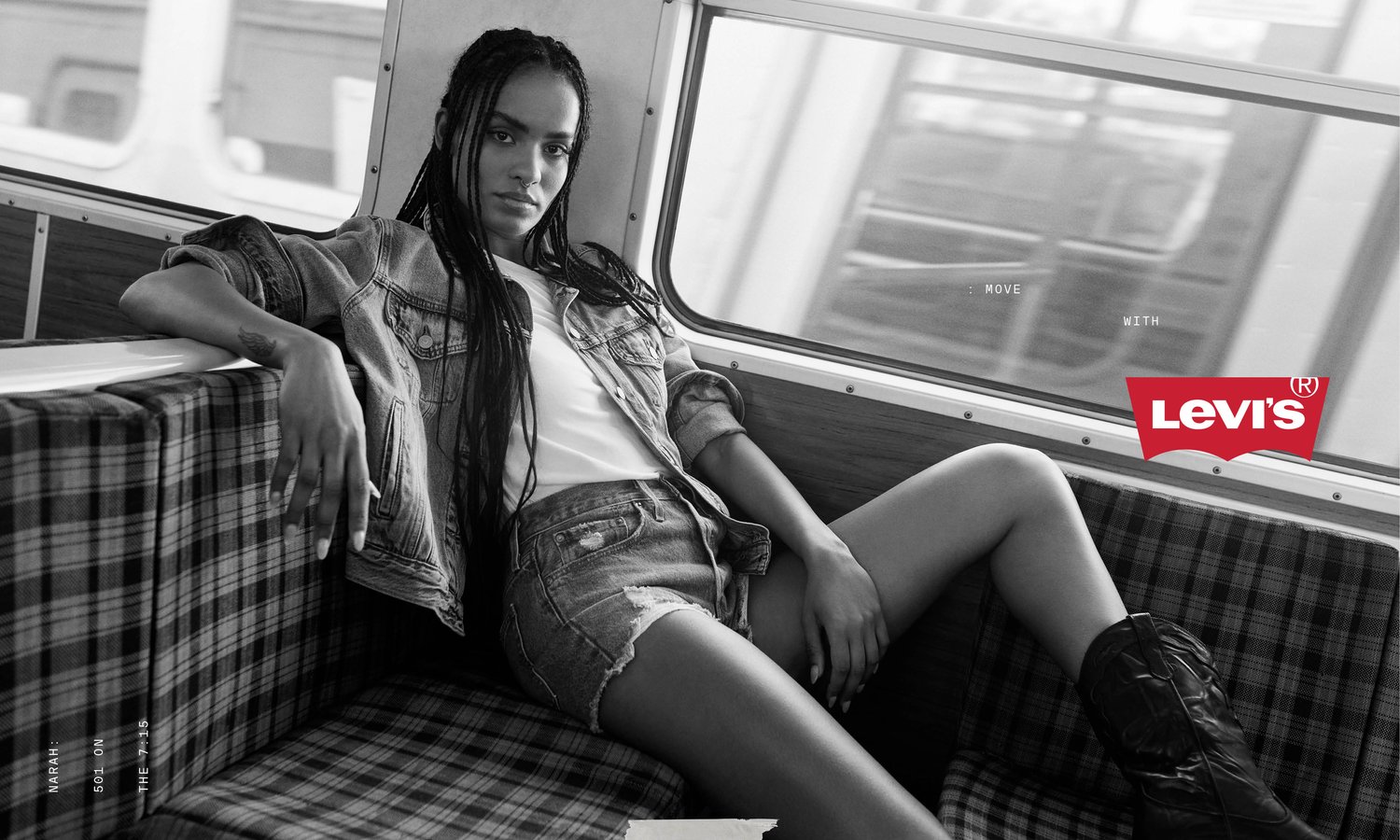 A woman in denim jacket and shorts sitting inside a train next to a window and wearing cowboy boots; black and white fashion shot.