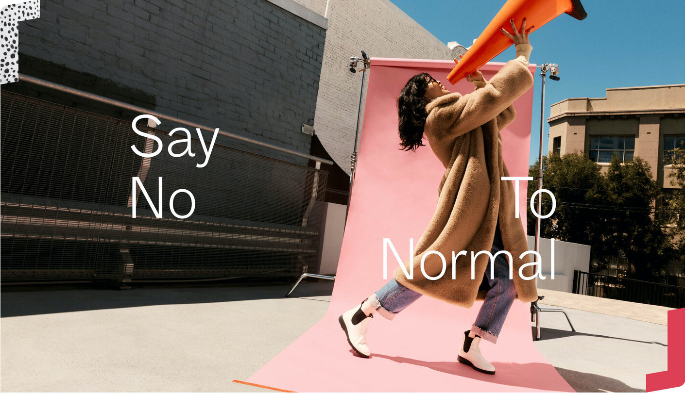 A woman in a tan coat uses a traffic cone as a megaphone against a pink backdrop, with the text 'Say No To Normal' overlaid.