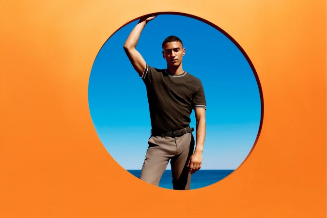 Man wearing a textured dark green t-shirt and light trousers posing inside a circular cutout against an orange wall.