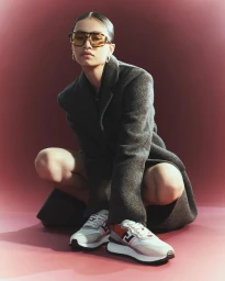 Model in oversized grey coat and tinted sunglasses squatting against a muted red gradient backdrop wearing patterned sneakers