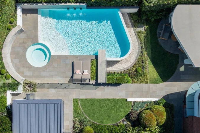 Overhead shot of a luxury backyard featuring a swimming pool, hot tub, lounge chairs, a stone patio, green landscaping, and modern architecture.