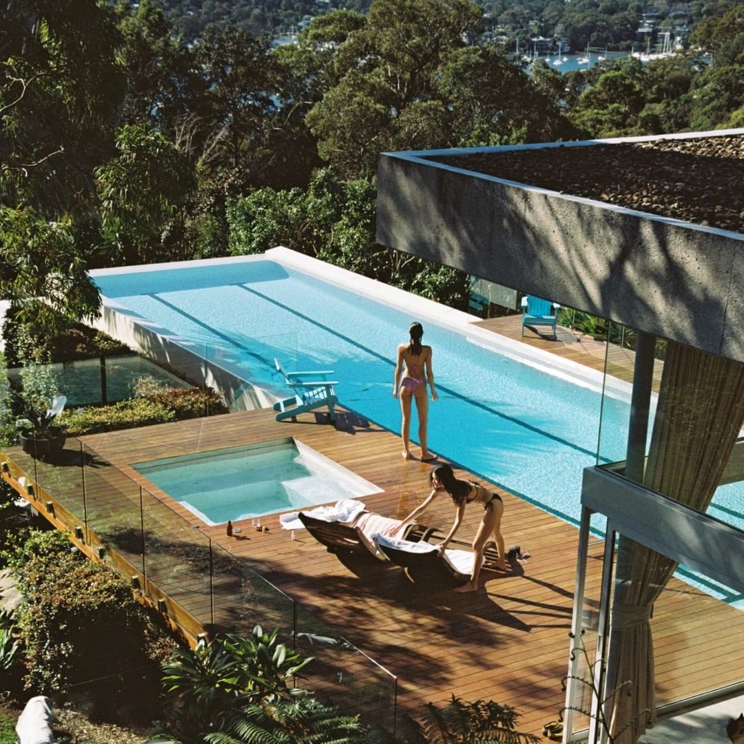 Two women relaxing by a luxury pool overlooking a green landscape. One woman is reclining on a lounger and another is attending to her.